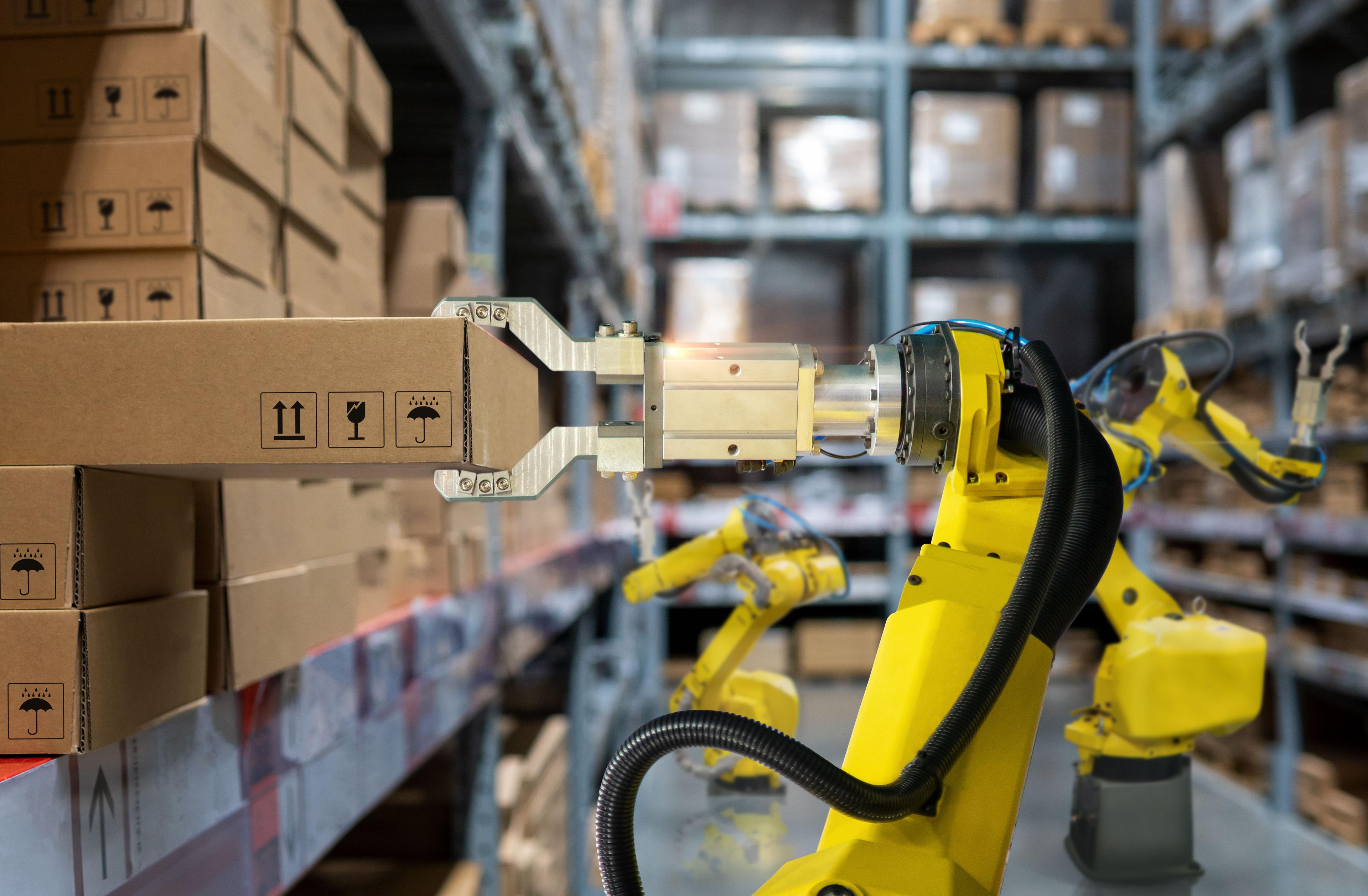 Yellow industrial robot in an automated warehouse sorting packages against the backdrop of tall shelves filled with cardboard boxes. The image illustrates modern AI and robotics solutions in re-commerce logistics, as discussed in the JLL material.
