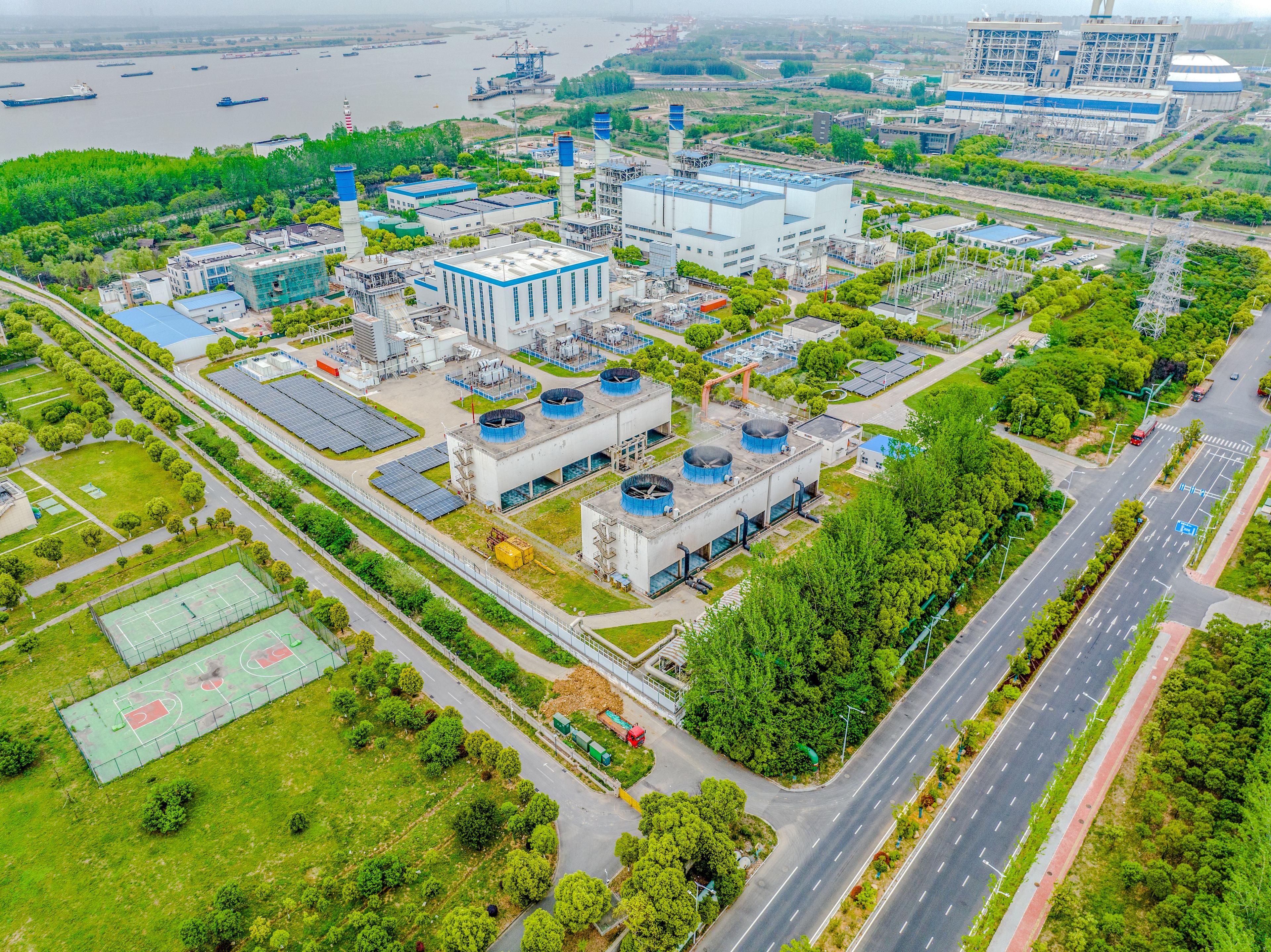 Aerial view of a modern warehouse with solar panels, and energy infrastructure surrounded by greenery, roads, and a sports court, located near a river with ships passing by.