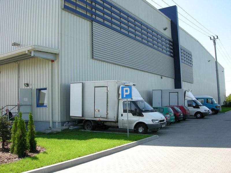 Zdjęcie prezentuje samochody na parkingu Logistics Center Kraków II, w tle fragment budynku magazynowego.