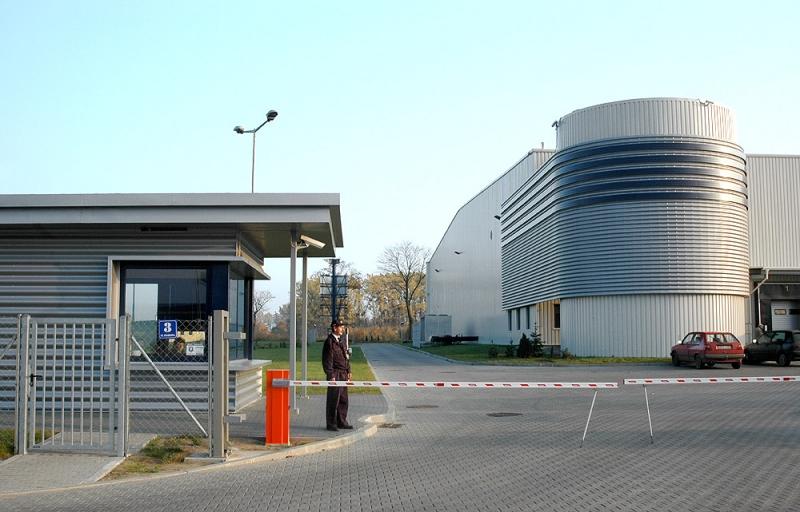 Zdjęcie przedstawia szlaban wjazdowy oraz stróżówkę Logistic Center Pruszcz Gdański, w tle widoczny fragment budynku magazynowego.