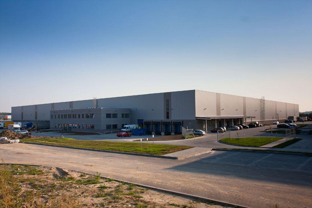 Zdjęcie prezentuje drogę dojazdową oraz teren Goodman Kraków Airport Logistics Centre, w tle obiekt magazynowy