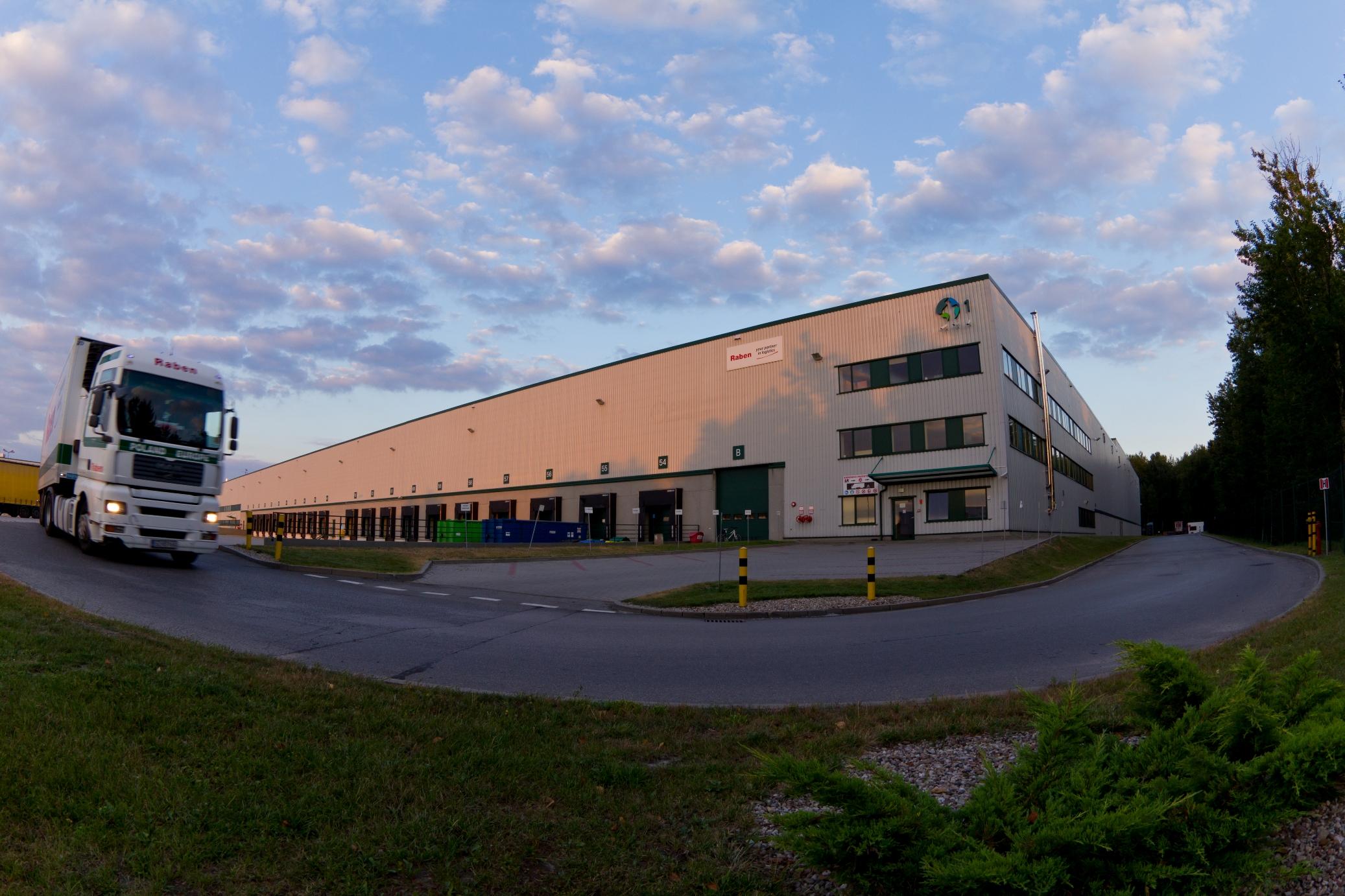Zdjęcie wykonane wieczorną porą, prezentuje nadjeżdzającą ciężarówkę, w tle parking oraz budynek magazynowy ''DC 1'' - Prologis Park Chorzów.