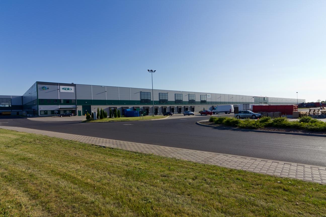 Zdjęcie przedstawia drogę dojazdową oraz parking wewnątrz kompleksu Prologis Park Wrocław III, w tle budynek magazynowy ''nr. 4'' .