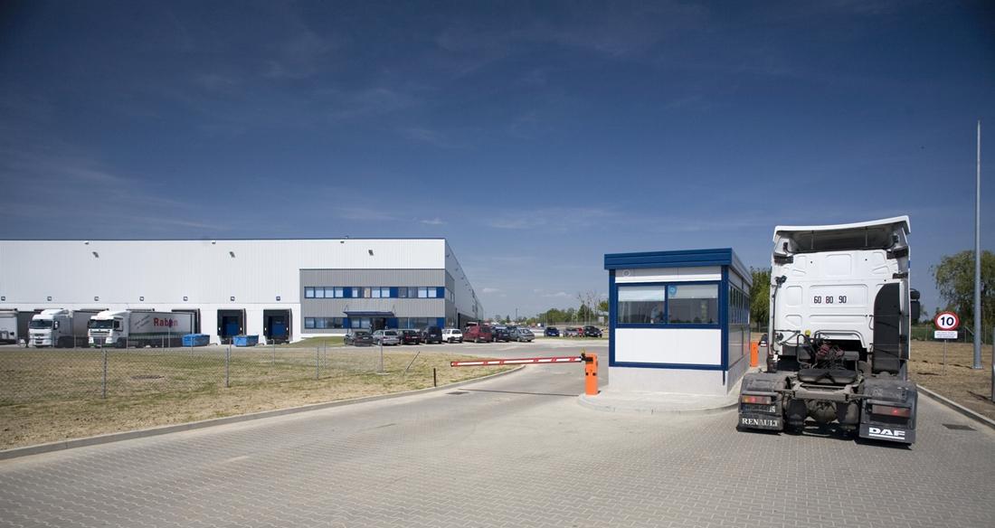 Zdjęcie przedstawia kontrolę ciężarówki, przed bramą wjazdową Ożarów Logistic Park, w tle parking oraz budynek magazynowo-biurowy.